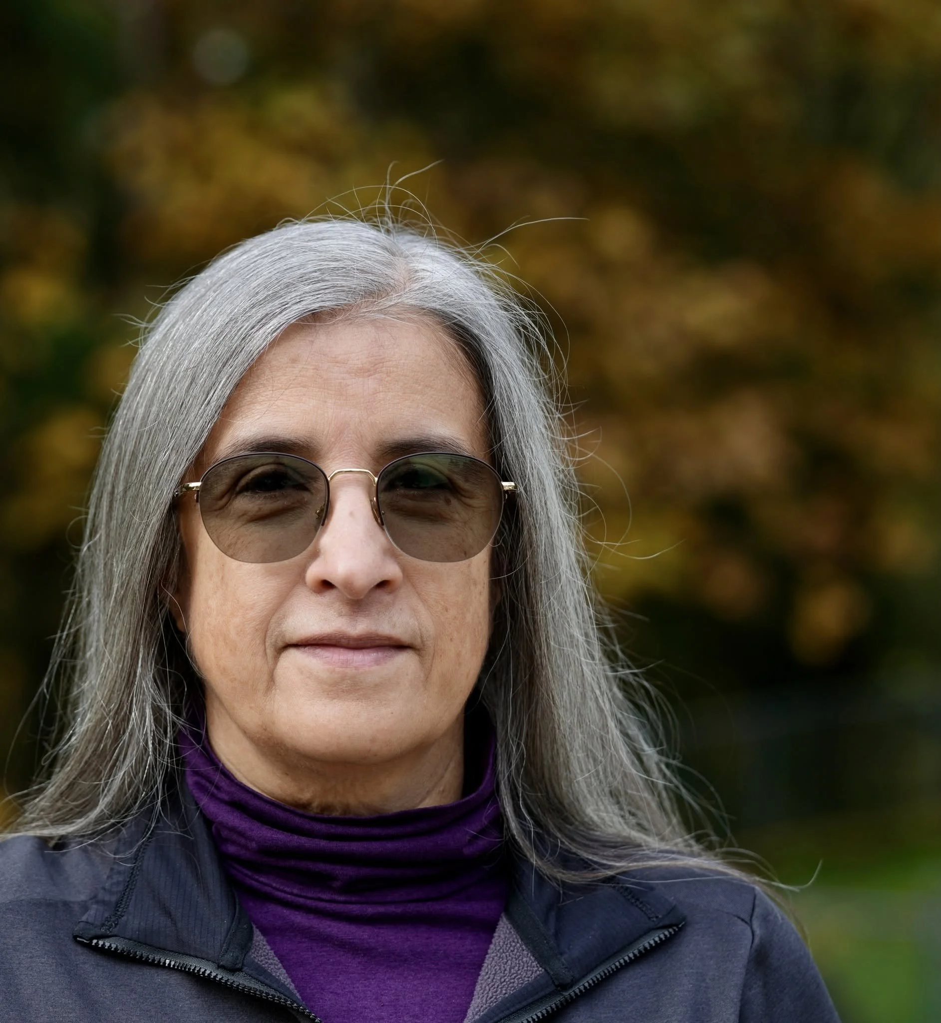 Close-up of an elderly woman with long silver hair wearing sunglasses, a purple turtleneck, and a dark jacket outdoors with a blurry background of trees.