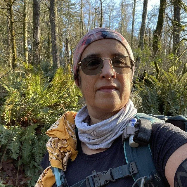 Woman wearing sunglasses, a headscarf, and a neck gaiter, taking a selfie in a forest with trees and ferns.