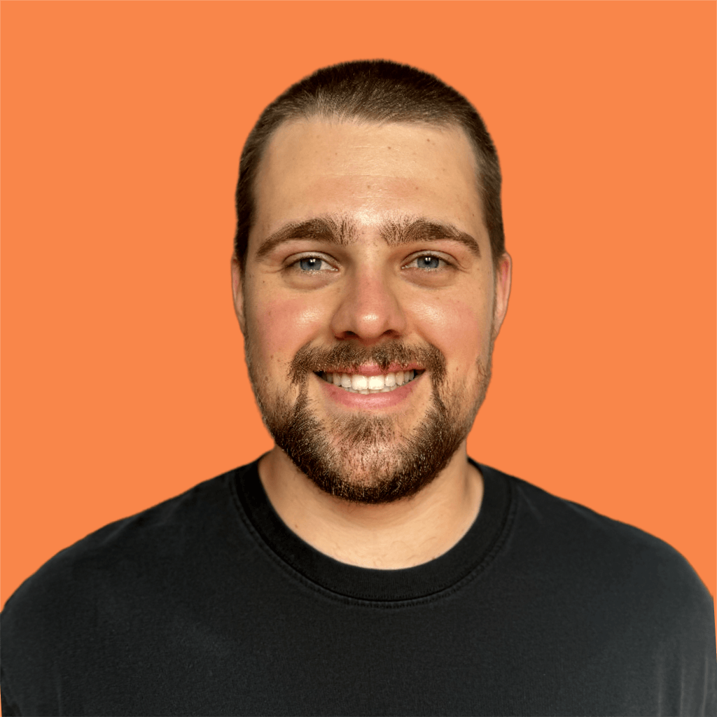 A young man with short brown hair and a beard, smiling, wearing a black t-shirt, against an orange background.