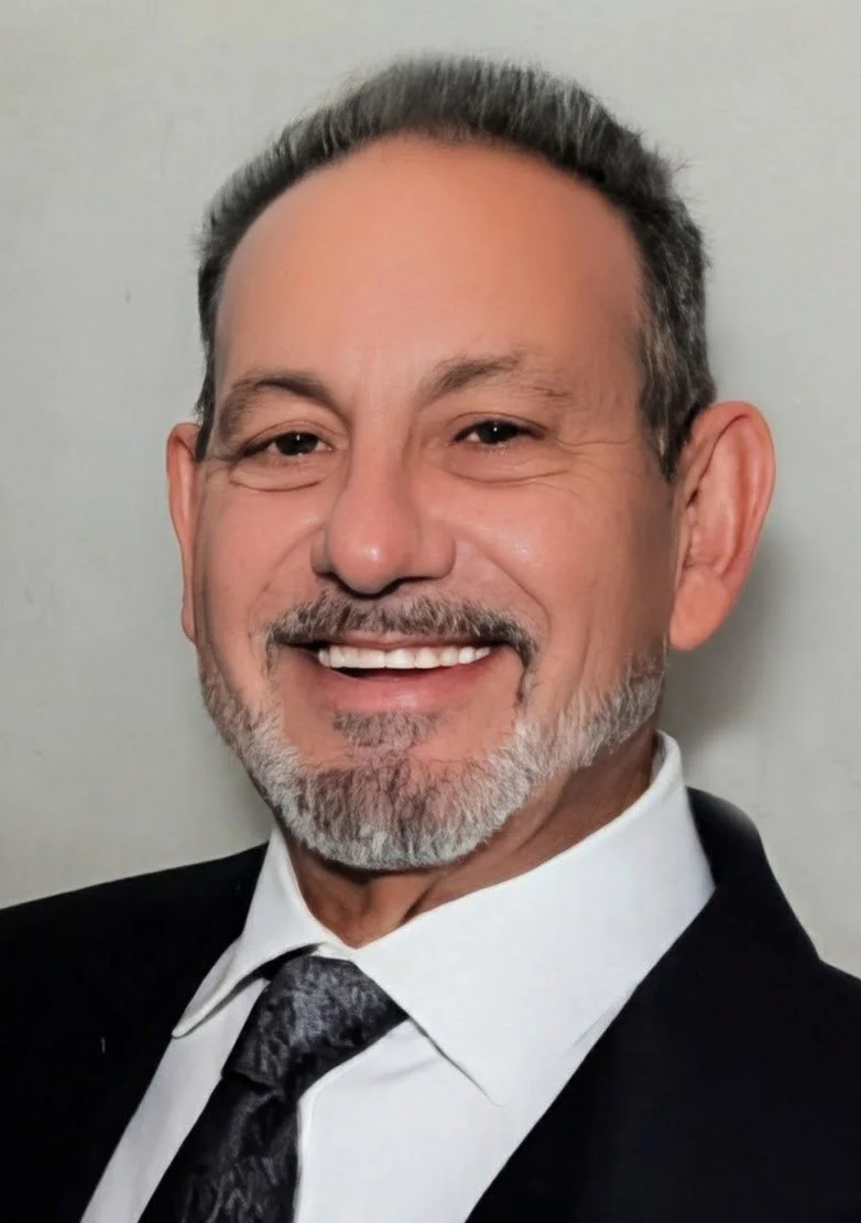 A middle-aged man smiling, dressed in a suit with a tie, standing against a plain background.