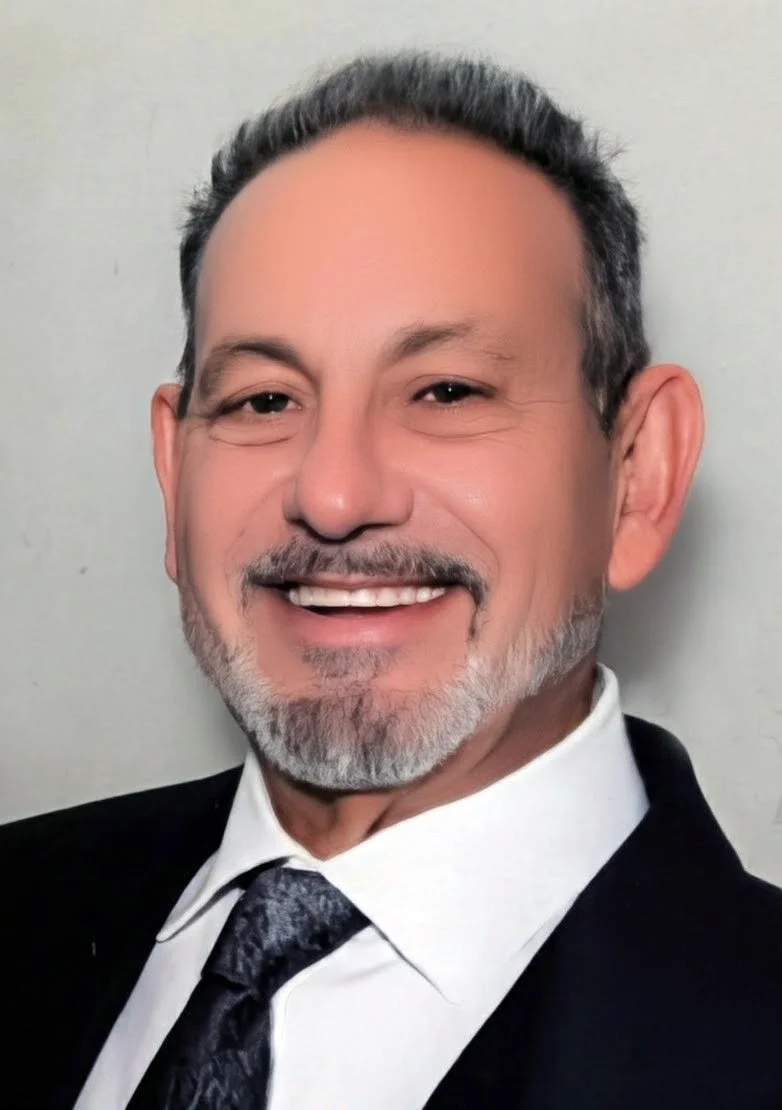 A smiling man with gray hair, beard, and mustache, wearing a black suit, white shirt, and gray tie, standing against a plain background.