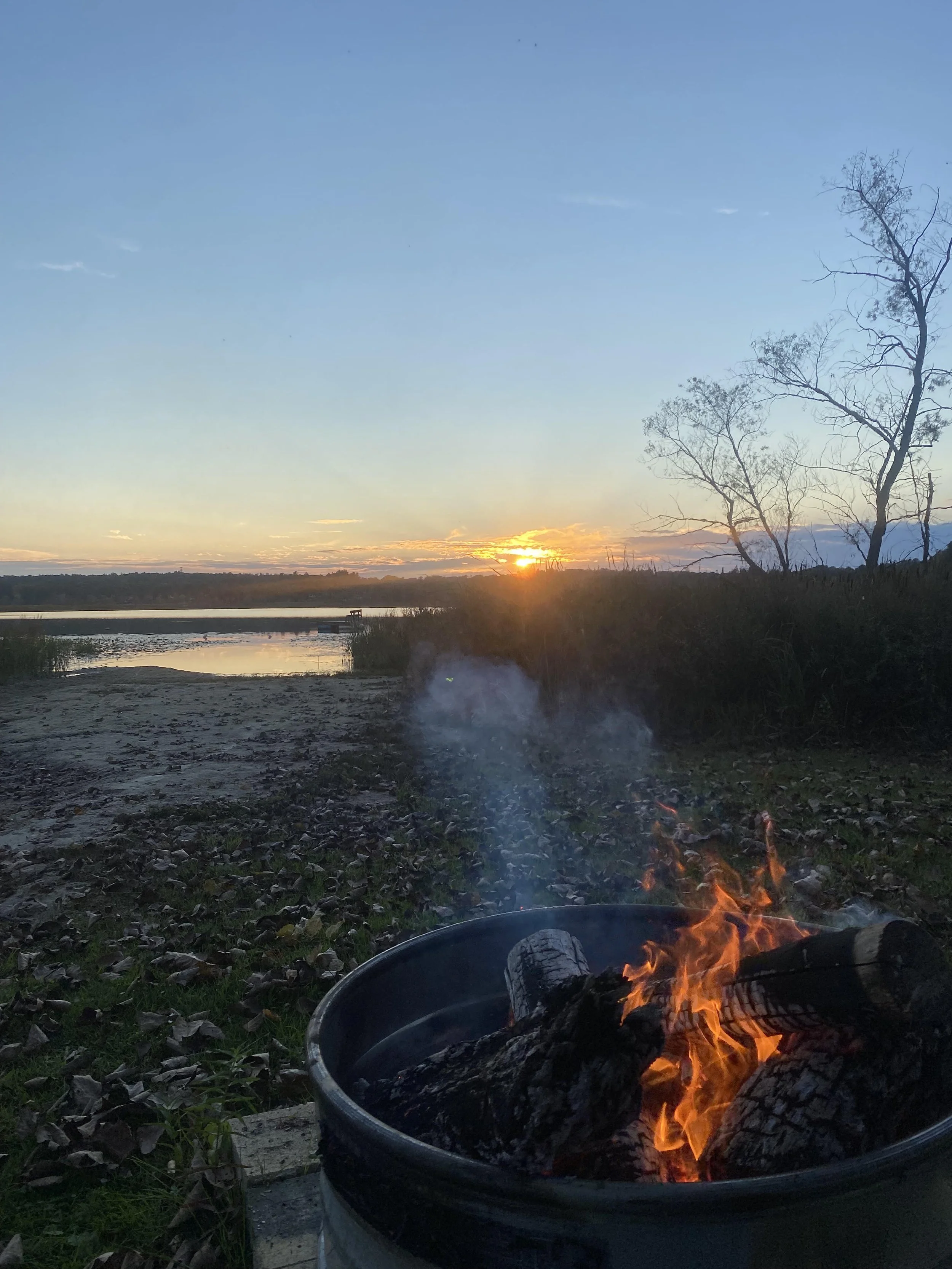View of a campfire burning logs at sunset near a river, with leafless trees and a clear sky in the background.