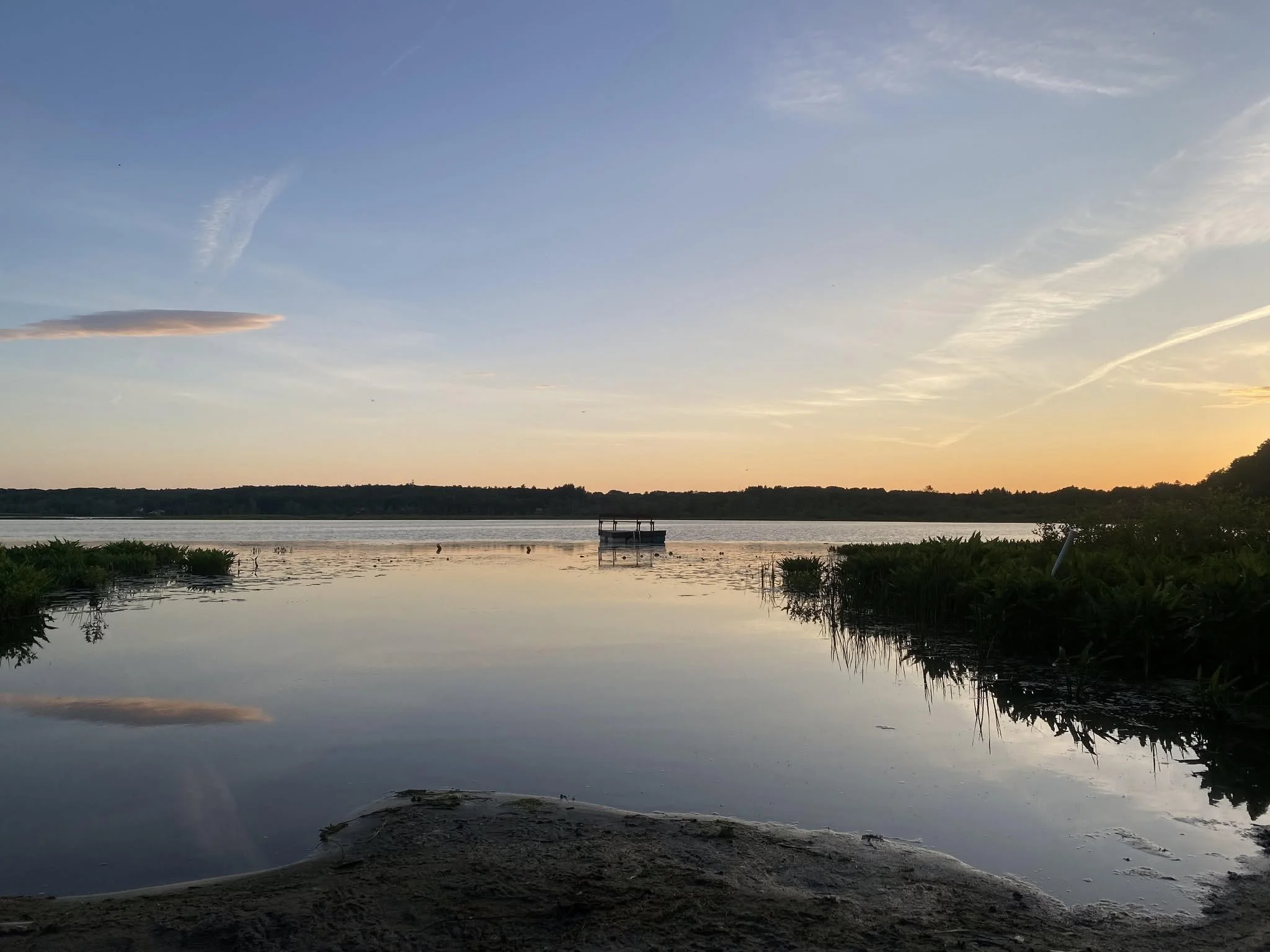 Scenic view of a calm lake at sunset with a small dock, trees along the shoreline, and a partly cloudy sky with pastel colors reflecting on the water.