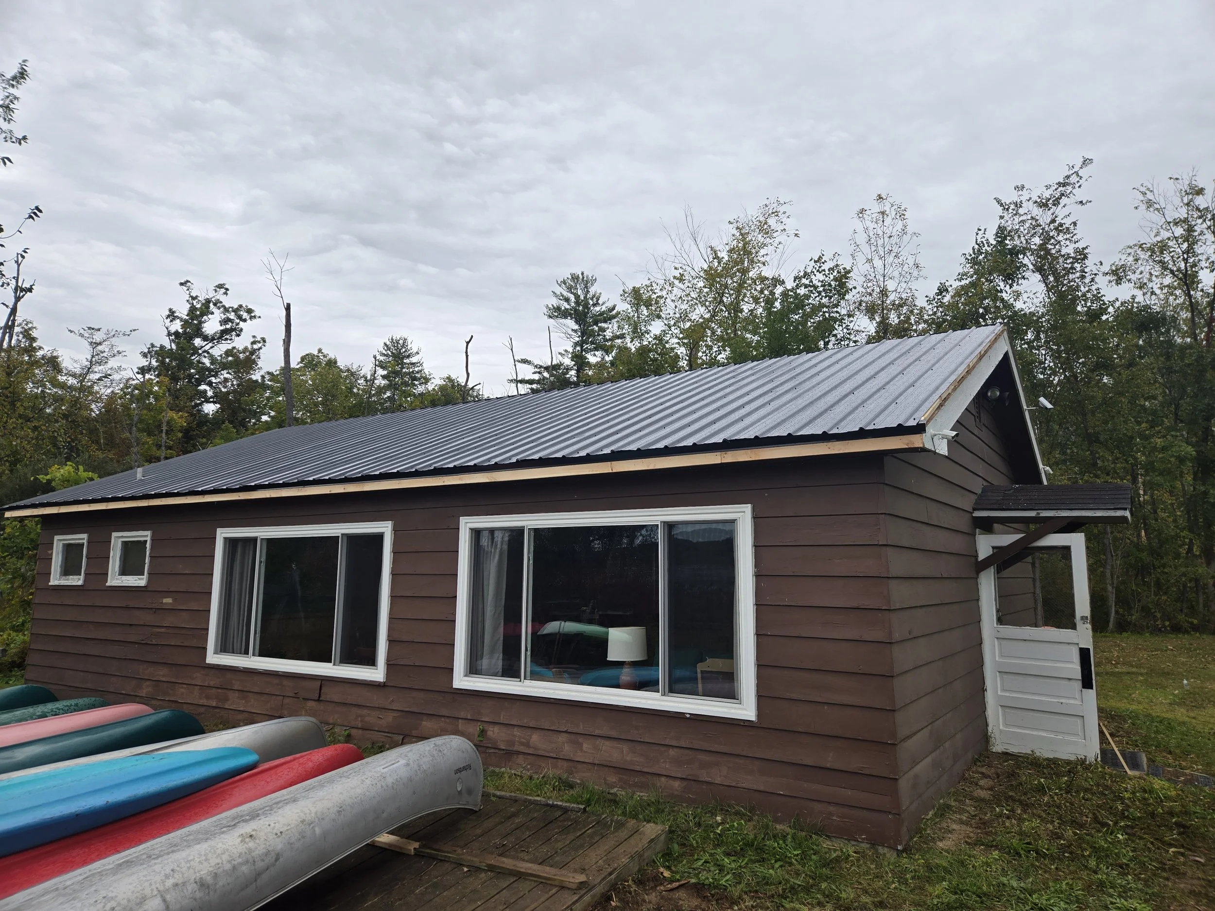 Exterior of a brown house with three large windows and a black metal roof. Several canoes are stacked on the wooden deck in front of the house, and a lush green wooded area is in the background under a cloudy sky.