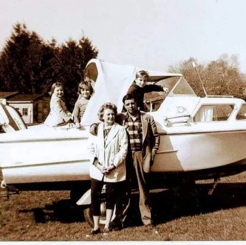 Vintage sepia family photograph of a mother and father standing in front of a small cabin cruiser on its trailer in a backyard, with three children sitting on and peeking out of the boat