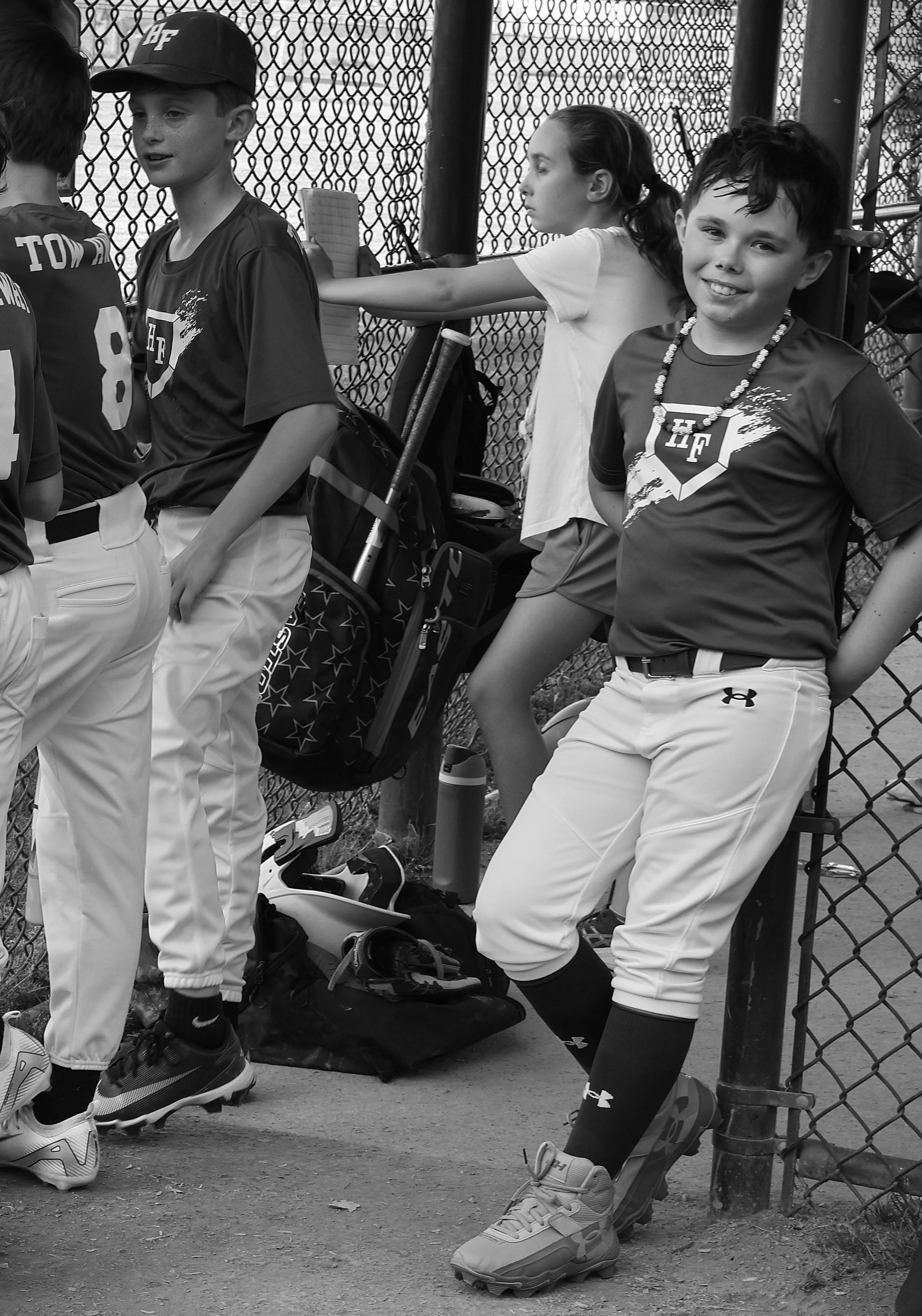 Black-and-white photograph of a young baseball player smiling toward the camera while teammates gather casually inside a chain-link dugout.
