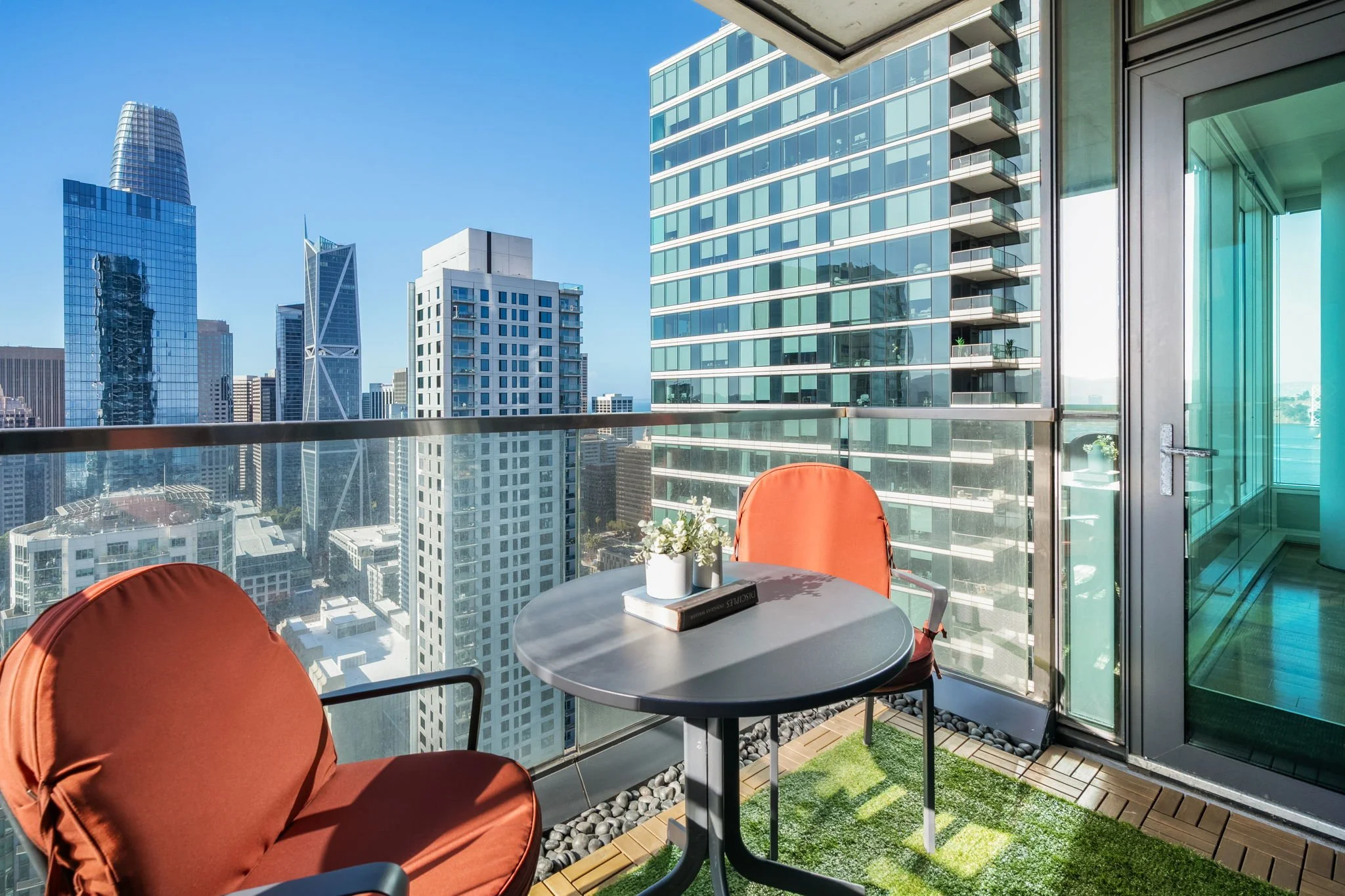 Balcony with round table, red chairs, potted plants, and view of city skyscrapers on clear day