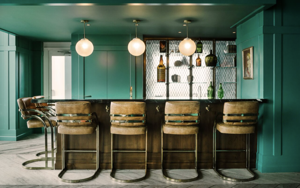 Interior view of a modern bar with teal walls, a black marble countertop, four upholstered bar stools, hanging globe pendant lights, and a back wall displaying bottles and decorations.