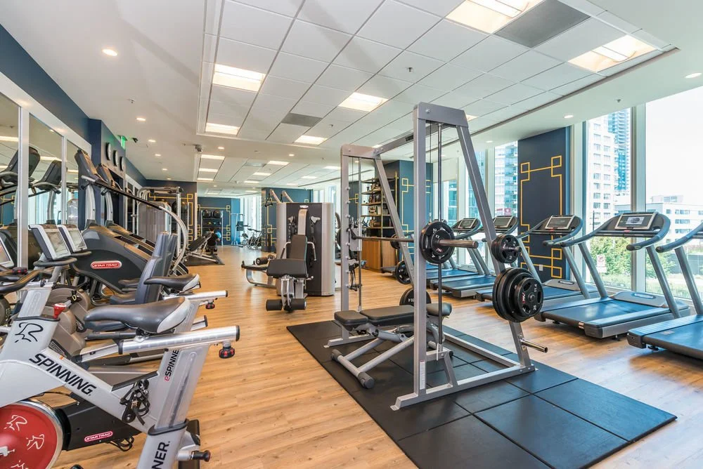 A modern gym with cardio equipment, including treadmills, elliptical machines, and a spinning bike, with large windows showing city buildings outside.