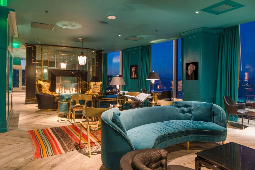 Elegant hotel lounge with teal velvet sofa, fireplace, various chairs, tables, lamps, large windows with city view, and artwork on teal walls.