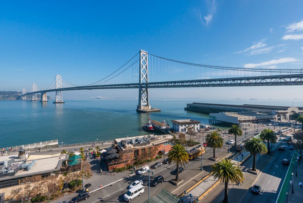View of the San Francisco-Oakland Bay Bridge over the water with a city street in the foreground, featuring palm trees and cars.