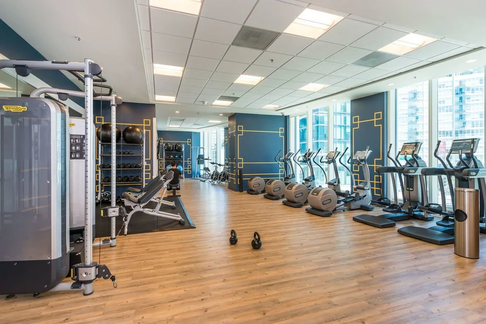 Empty fitness center with cardio machines, weightlifting equipment, kettlebells, and large windows showing cityscape outside.