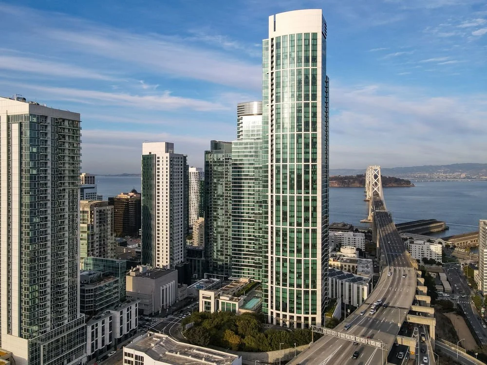 Skyscrapers and high-rise buildings in a city by the water, with a bridge connecting to another landmass, under a partly cloudy sky.