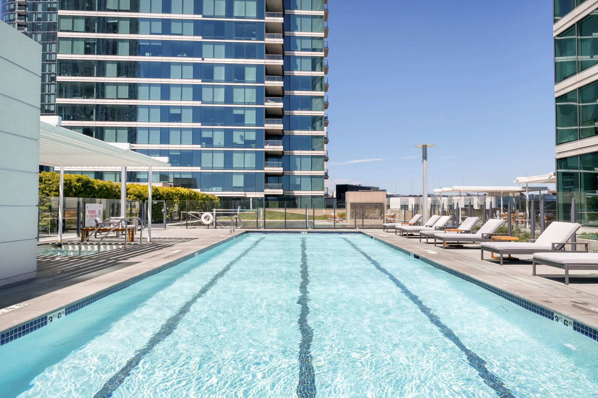 View of a rooftop swimming pool in a modern urban apartment complex, with lounge chairs, patio tables, and surrounding high-rise buildings under a clear blue sky.