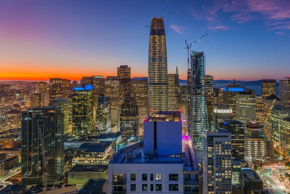 City skyline at sunset with tall skyscrapers and construction cranes.