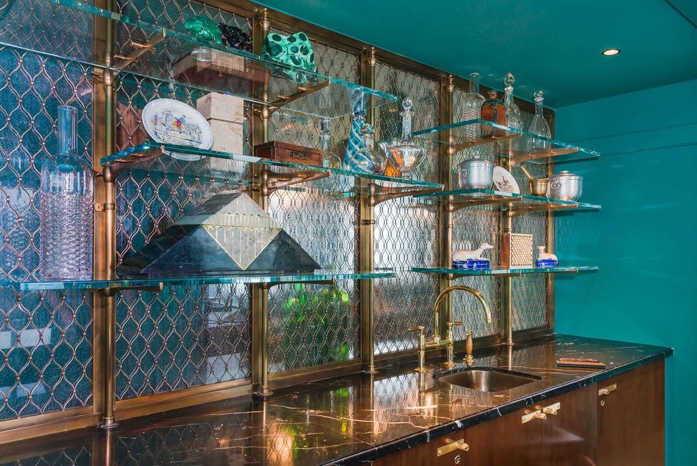 Open kitchen shelf with glass shelves, decorative bottles, ceramic dishes, and a gold faucet.