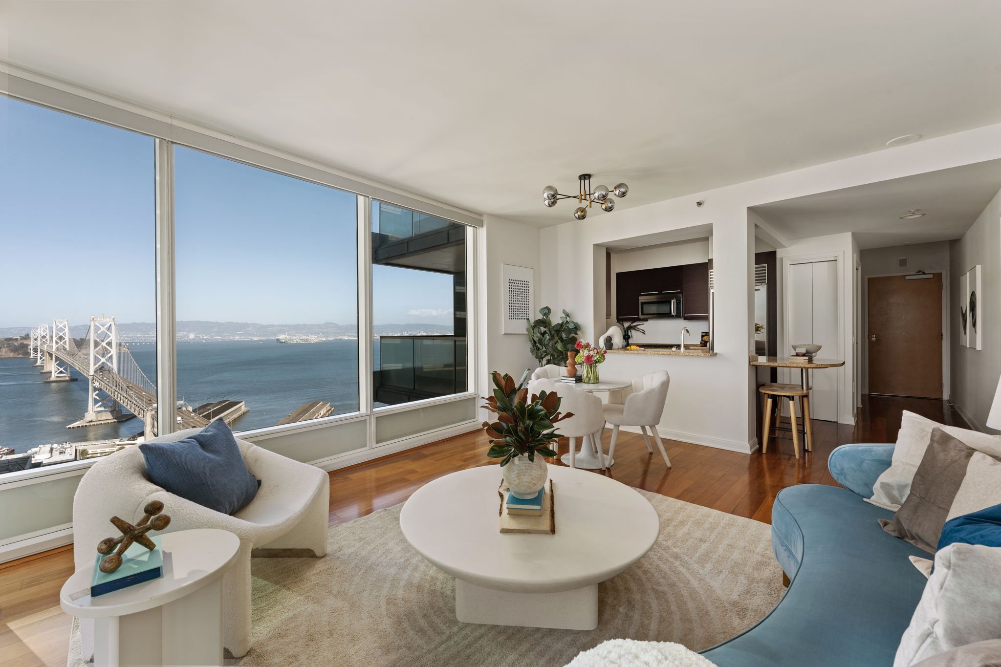 Modern living room with large windows showing a bridge and water view, featuring white and blue furniture, a round coffee table, indoor plants, and a kitchen area in the background.