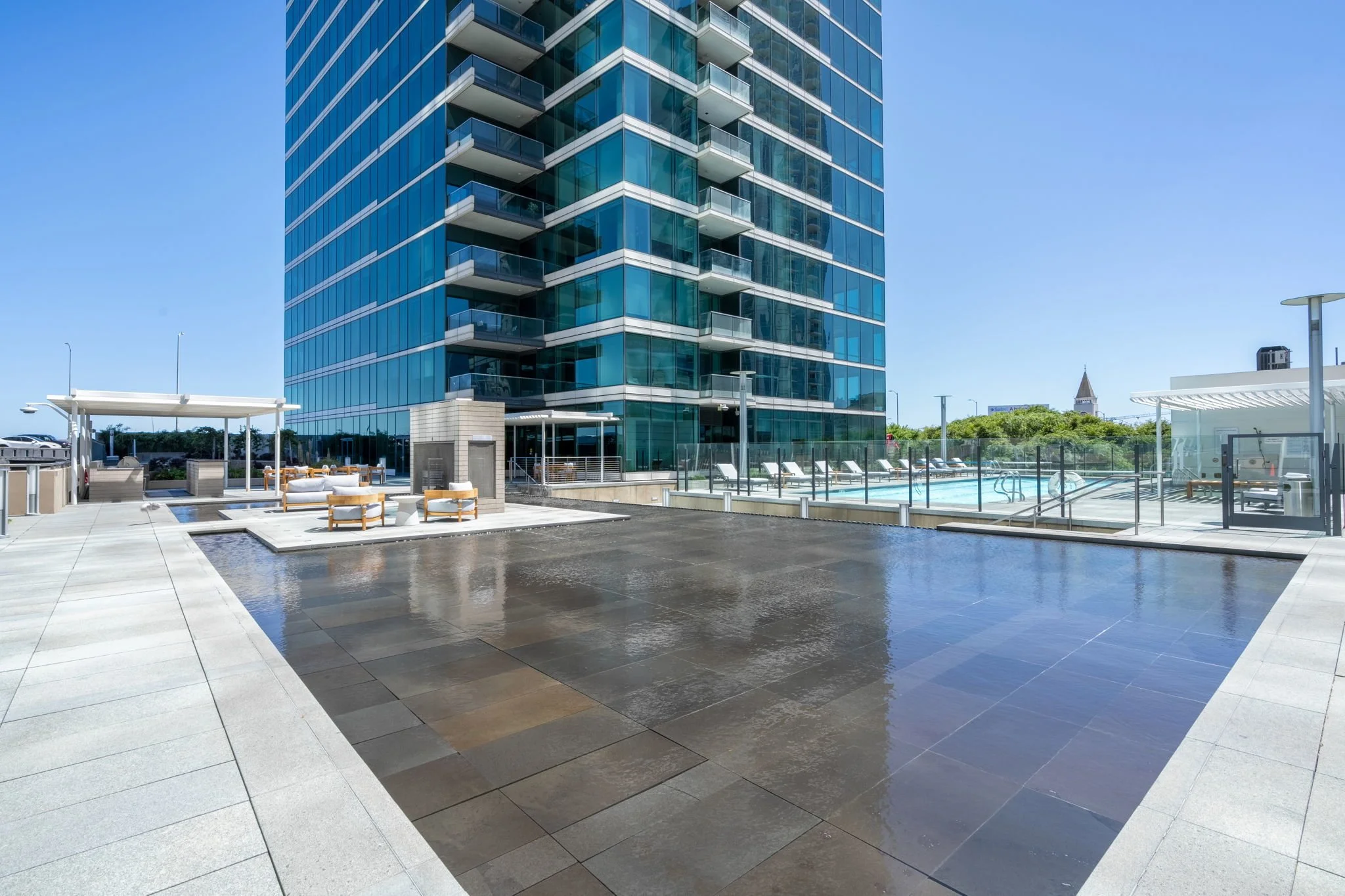 Modern high-rise building with glass windows and balconies, pool area with lounge chairs and shade structures, clear blue sky, cityscape in the background.