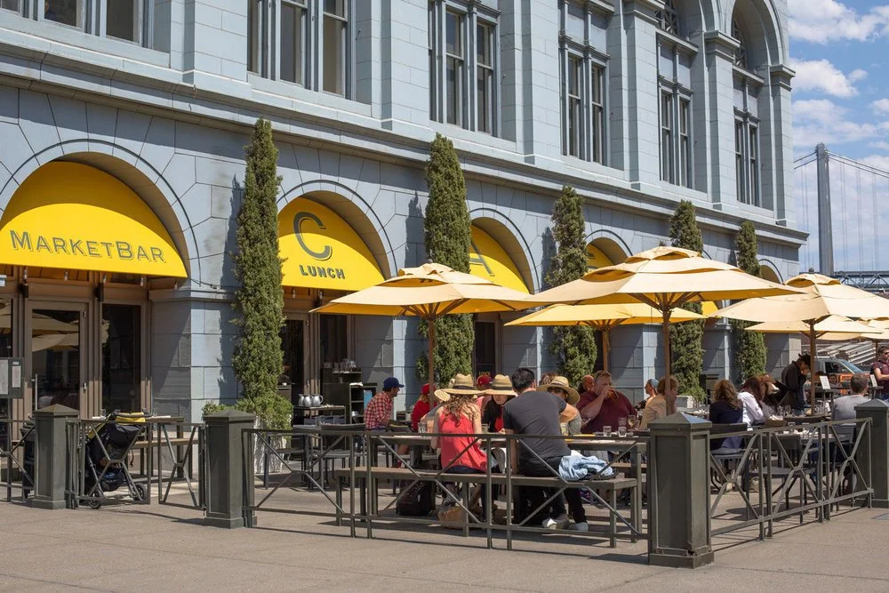 People dining outdoors at a restaurant with yellow umbrellas and yellow awnings on a sunny day, with a historic grey building and bridge in the background.