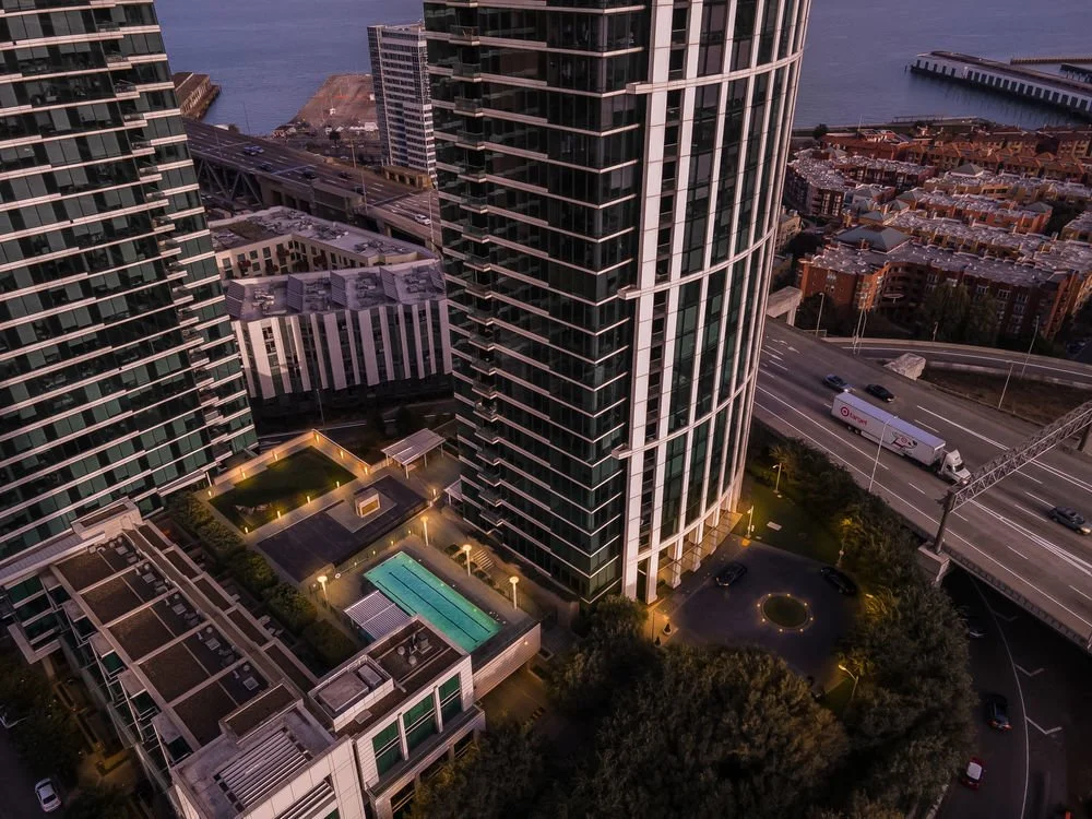 Aerial view of tall modern glass skyscrapers, a swimming pool, and surrounding cityscape near water, with roads and vehicles at dusk.