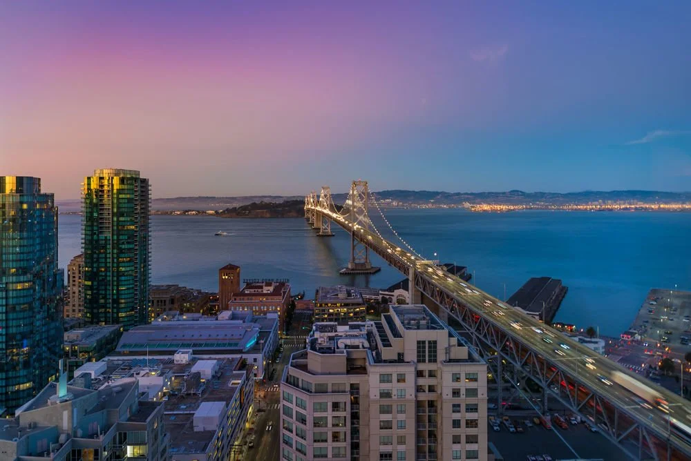 Cityscape of San Francisco at sunset with the Bay Bridge over the water, tall buildings, and a colorful sky.