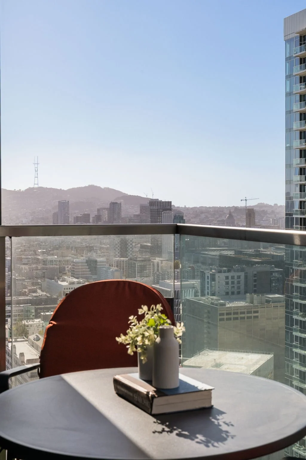 A balcony with a round table, a brown chair, a white vase with flowers, and a city skyline view of tall buildings and hills in the distance under a clear blue sky.