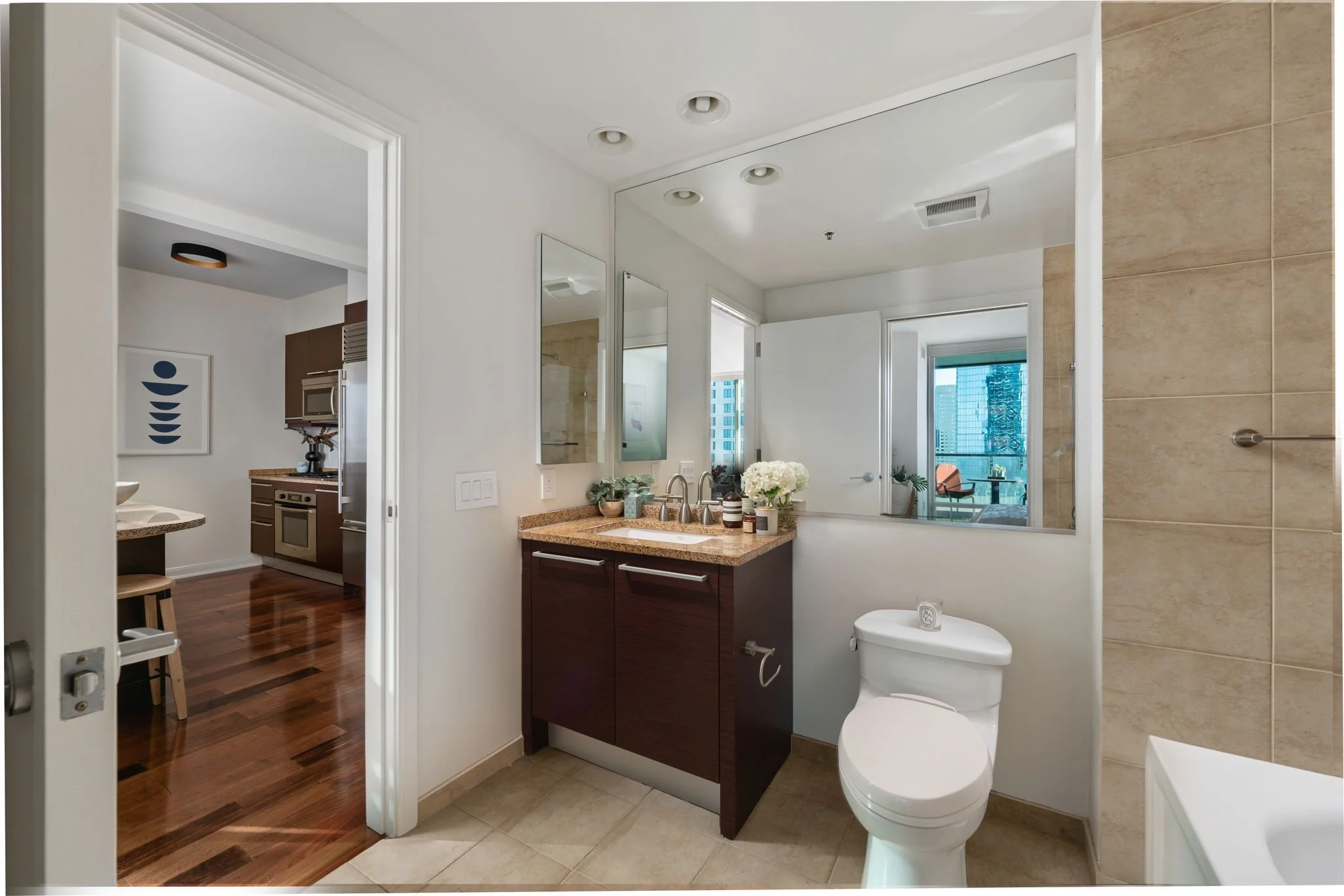 Modern bathroom with a white toilet, dark wood vanity, granite countertop, mirror, and beige tiled wall, with a view into a balcony through a window.
