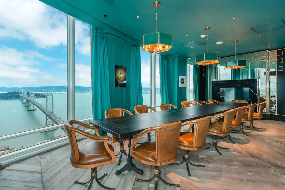 A modern conference room with a long black table, tan leather chairs, large windows overlooking a bridge and water, teal walls and curtains, and pendant lights.