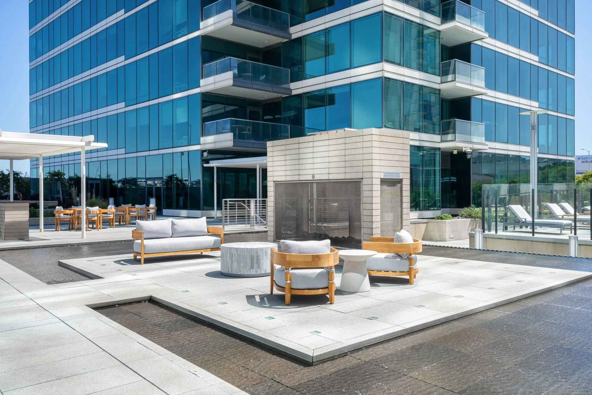 Outdoor patio area with modern wooden and concrete furniture, glass building with balconies, and a small water feature.