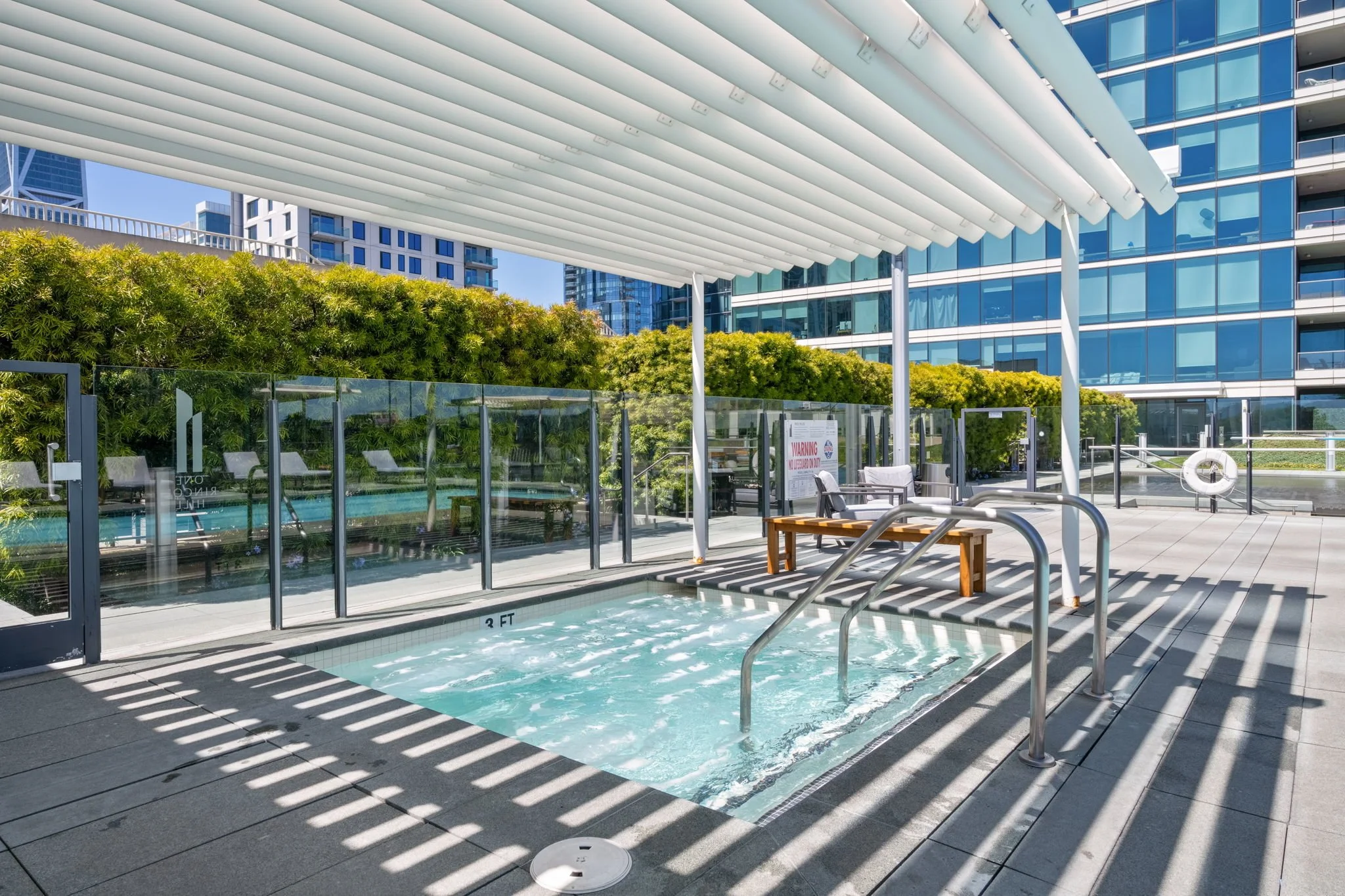 Outdoor pool area with a hot tub, shaded by a modern white canopy, with high-rise buildings and greenery in the background.