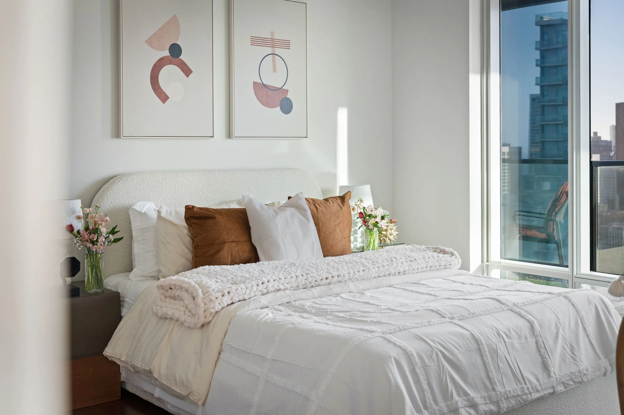 A bedroom with a bed, brown pillows, a white fuzzy blanket, and two vases of pink flowers on bedside tables. Two abstract art pieces hang above the bed. A large window shows a city view with tall buildings.