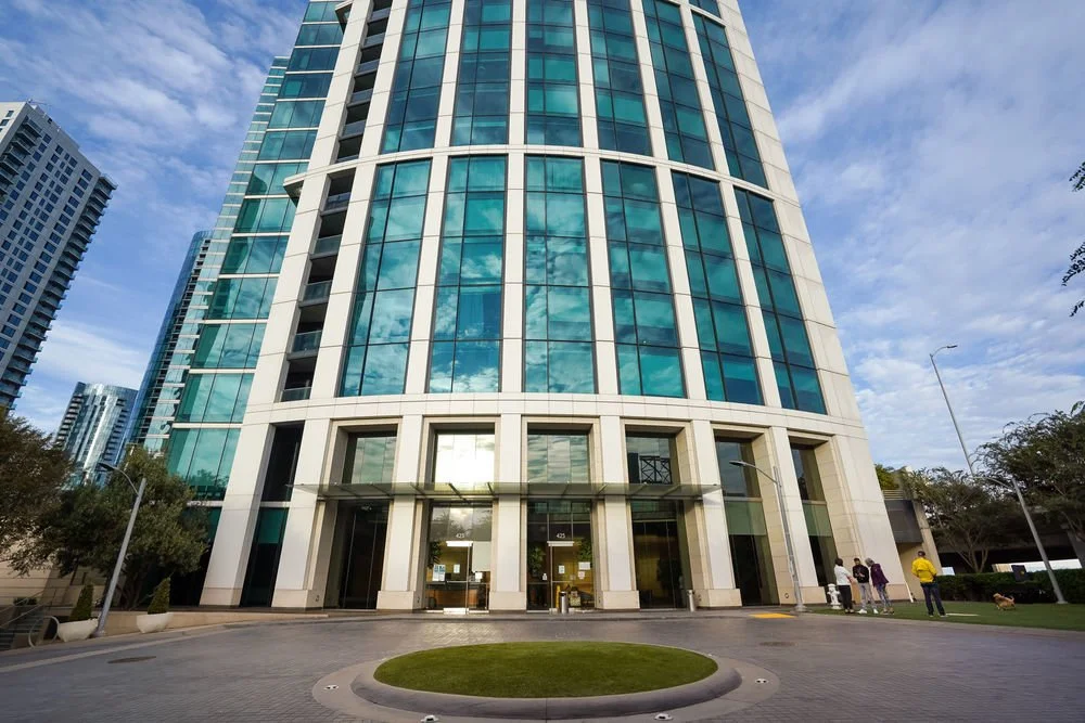 Tall modern office building with glass windows reflecting the sky and clouds, with a few people standing near the entrance and trees around.