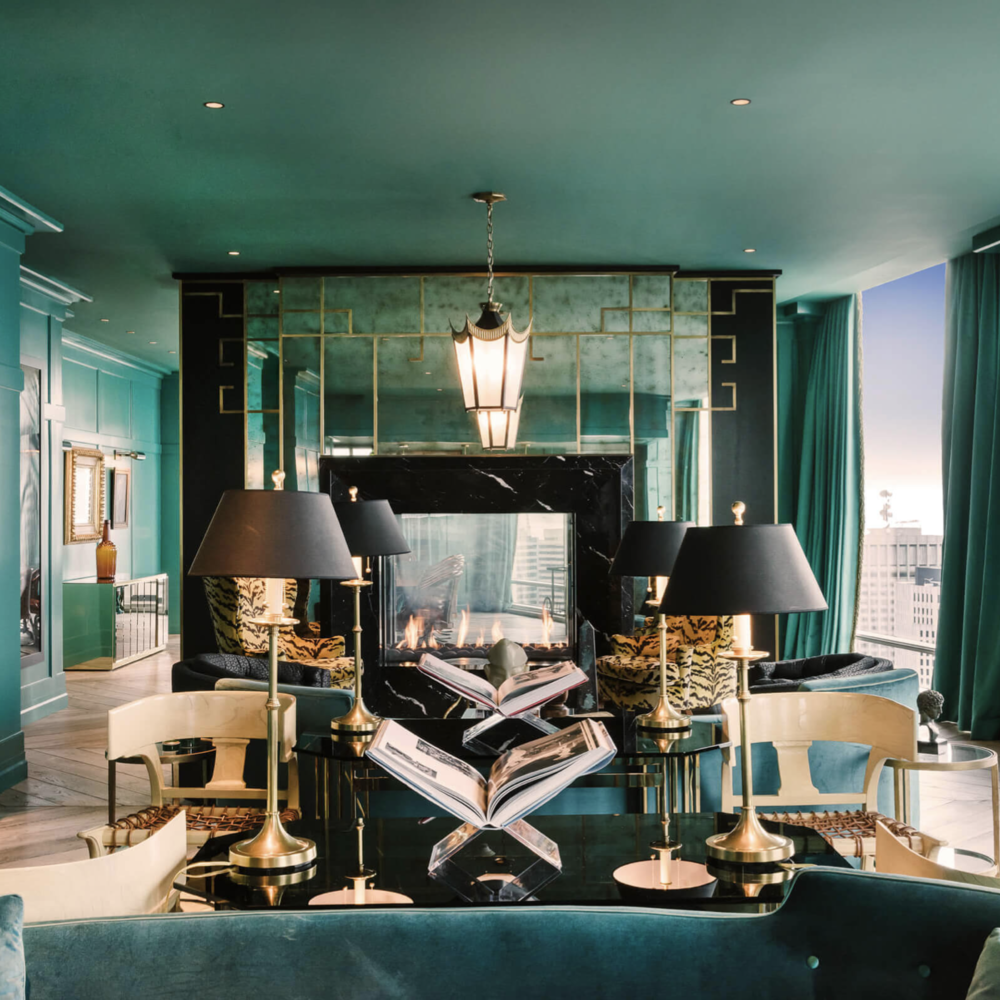 Living room with teal walls, black fireplace, large windows with teal curtains, and a mirror above the fireplace. There are black lamps with gold bases on the table, and books open on the table. The room has a mix of modern and classic decor.