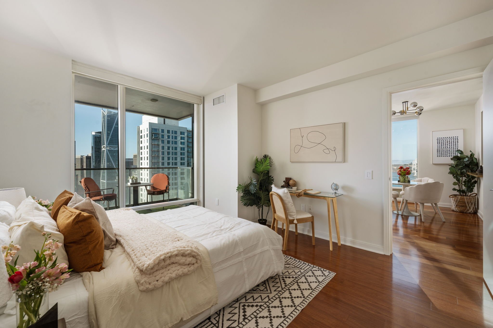 A cozy bedroom with a large bed covered in white bedding and pillows, adjacent to a balcony with city view through large windows, hardwood floors, and a small desk with a chair. The living room visible through an open doorway has a dining table, plan