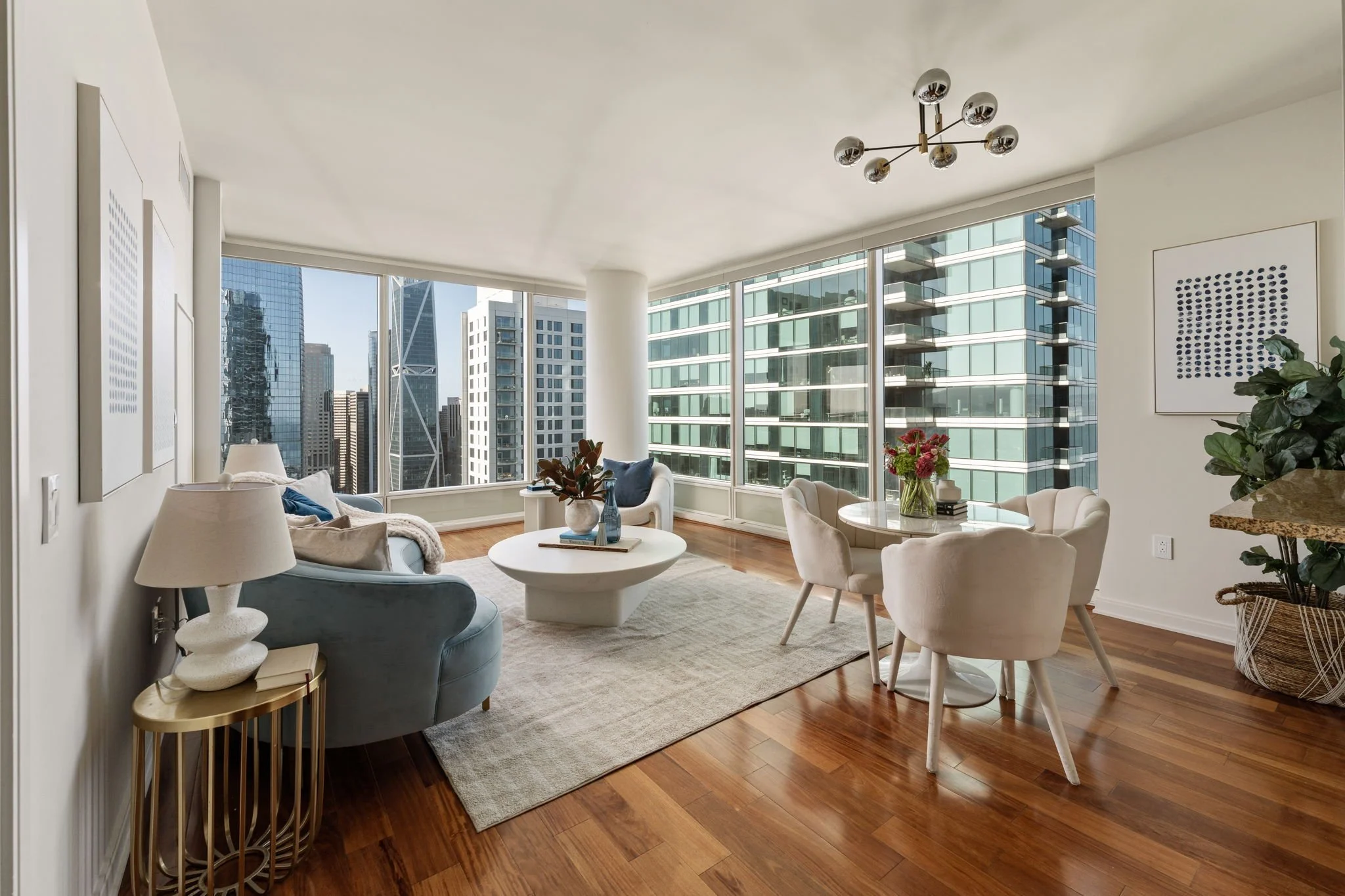 Modern living room with large windows, city skyline views, white and beige furniture, decorative plants, artwork, and hardwood floors.