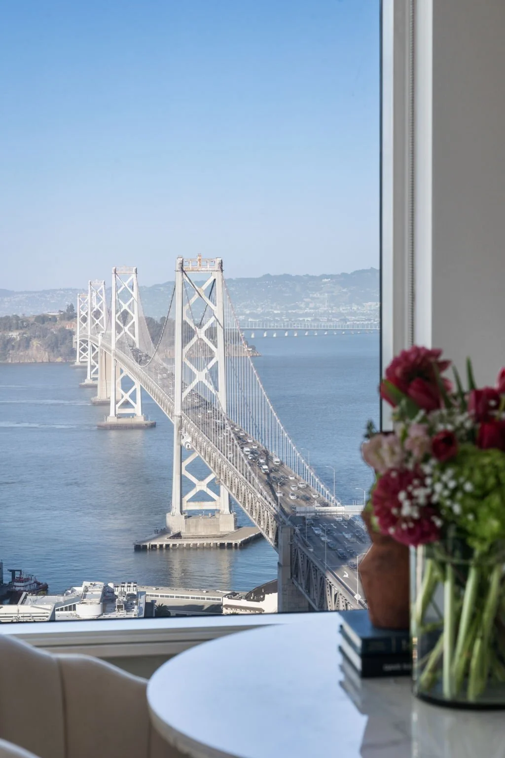 View of the San Francisco-Oakland Bay Bridge from inside a room, with a vase of flowers on a table in the foreground.