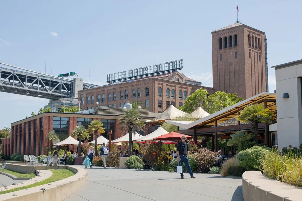 Outdoor shopping and dining area with people walking and sitting among umbrellas and trees, with a large brick building labeled 'Hills Bros Coffee' and a tall tower behind.