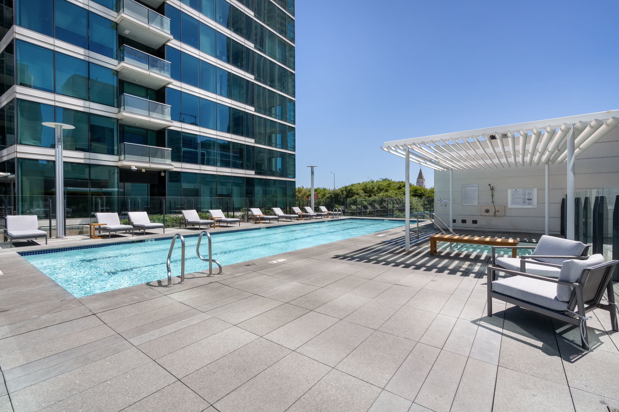 Empty outdoor swimming pool area on a sunny day, with lounge chairs, a white pergola, and a modern glass apartment building in the background.