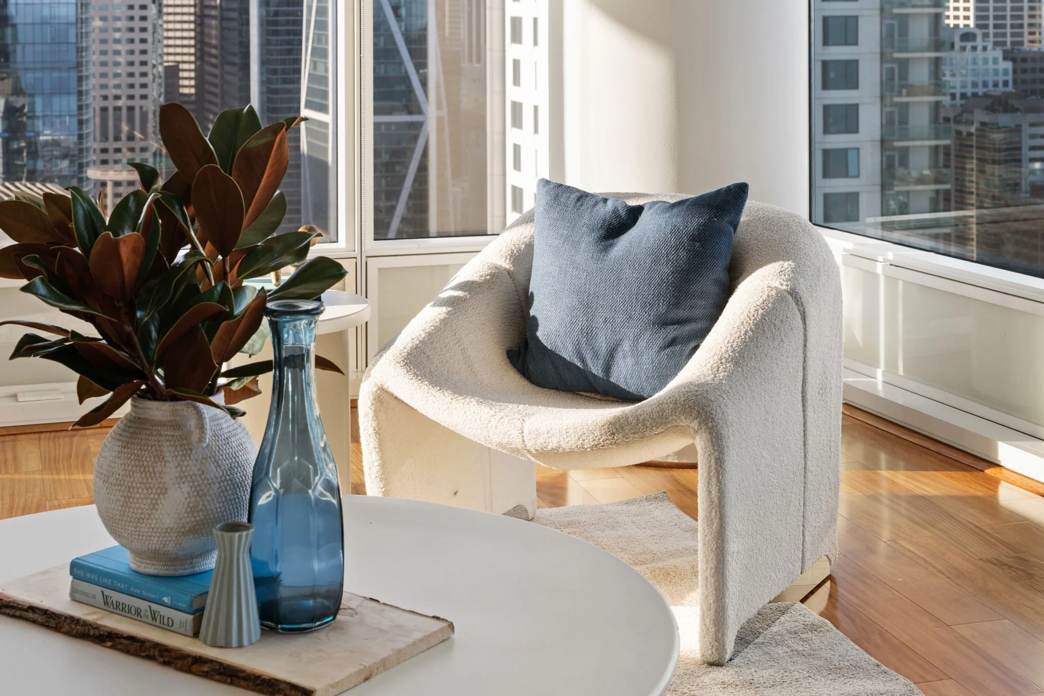 A cozy sitting area in a high-rise apartment with large windows showing cityscape. There is a white armchair with a blue pillow, a wooden table with a ceramic vase, blue bottles, and books, and a leafy potted plant.