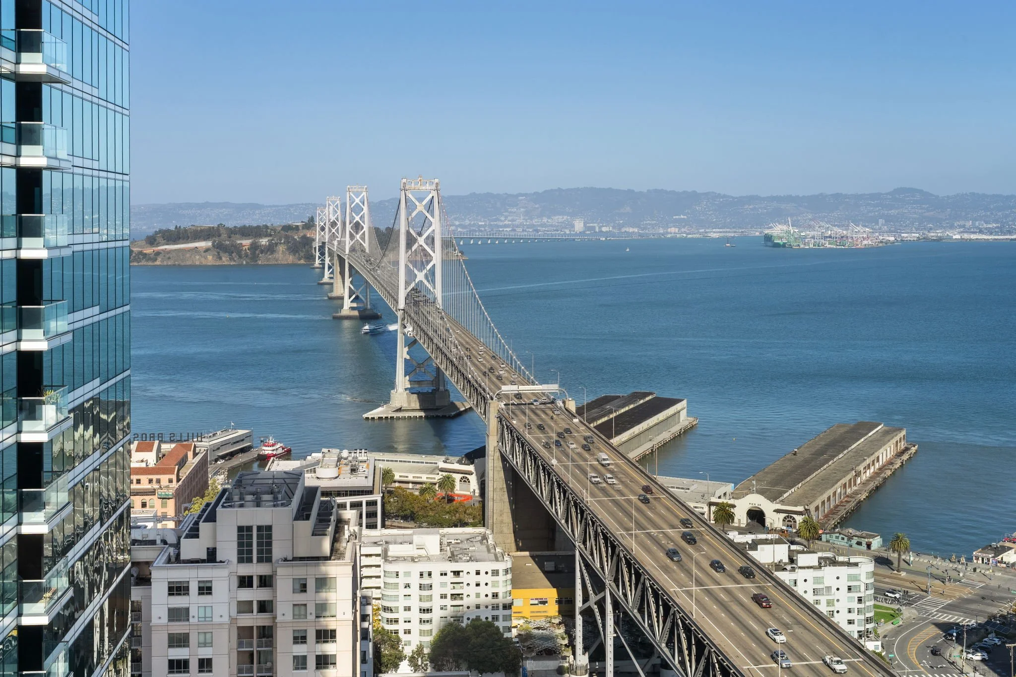 View of the San Francisco-Oakland Bay Bridge with city buildings in the foreground and water in the background.