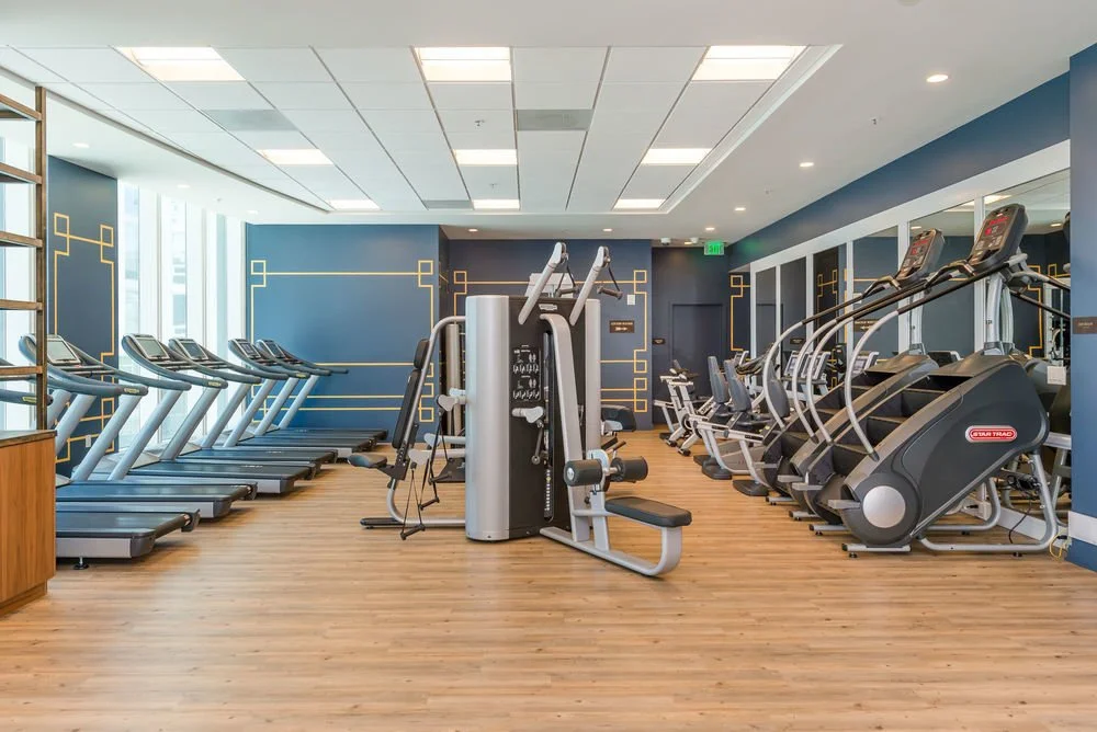 Empty gym room with treadmills, elliptical machines, and other fitness equipment on wooden flooring, with blue and white walls.