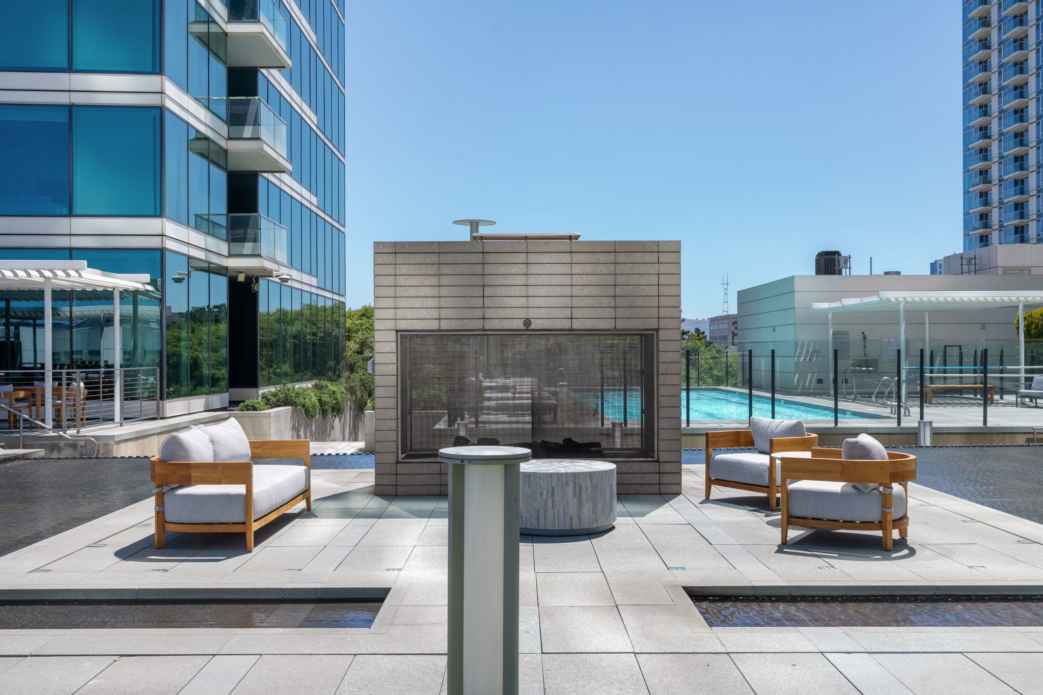 An outdoor rooftop area with modern lounge chairs, a small round table, a fireplace, and a pool with a glass safety fence. Surrounding skyscrapers reflect on the glass buildings and blue sky.