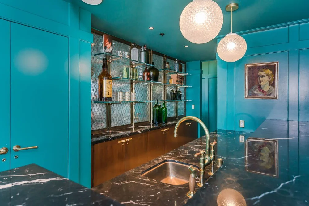 Modern home bar with turquoise walls, black marble countertop, brass fixtures, glass bottles on metal shelves, and a framed portrait of a woman on the wall.