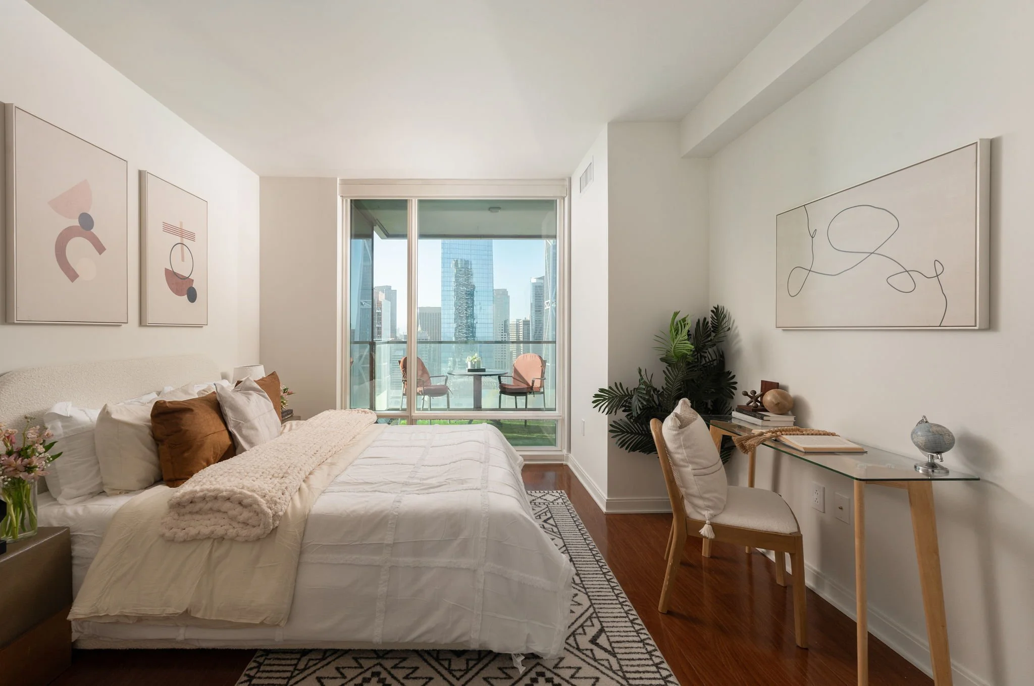 A modern bedroom with white walls, a large window overlooking a cityscape, a bed with white bedding and brown pillows, abstract artwork on the walls, a small desk with a glass top, a chair, and green plants.