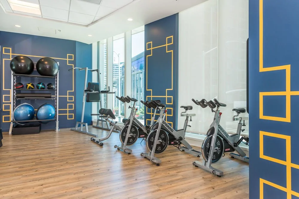 Fitness room with stationary bikes, exercise balls, and a pull-up station, featuring large windows and blue and yellow wall accents.