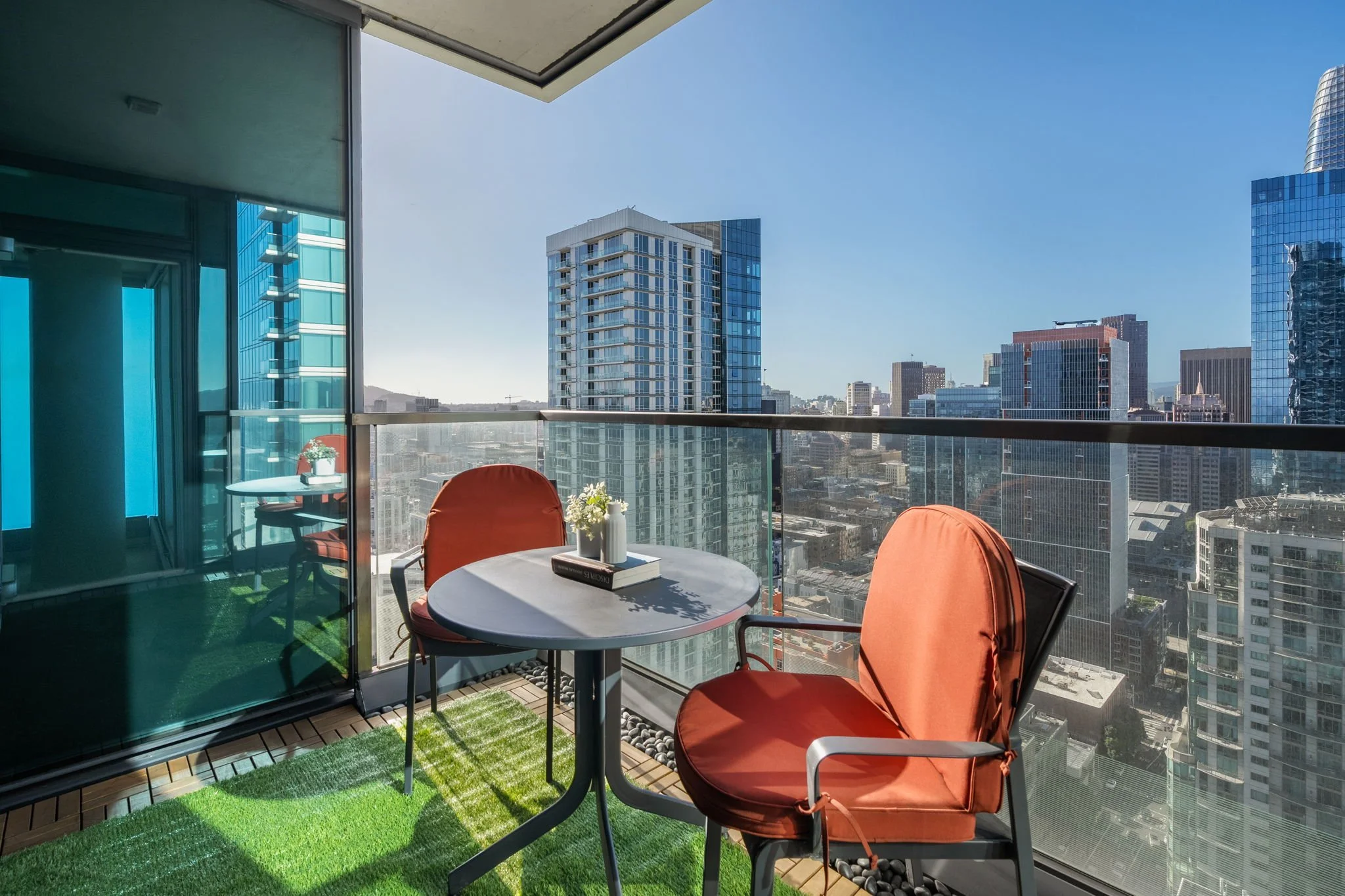 Balcony with round table, orange chairs, and view of a city skyline with tall modern glass buildings under a clear blue sky.