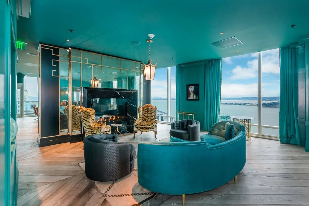Living room with teal curtains, a fireplace, and a view of a body of water through large windows. The room features a mix of modern and vintage furniture, including a curved blue sofa, two tiger-patterned armchairs, and small side tables. A chandelie