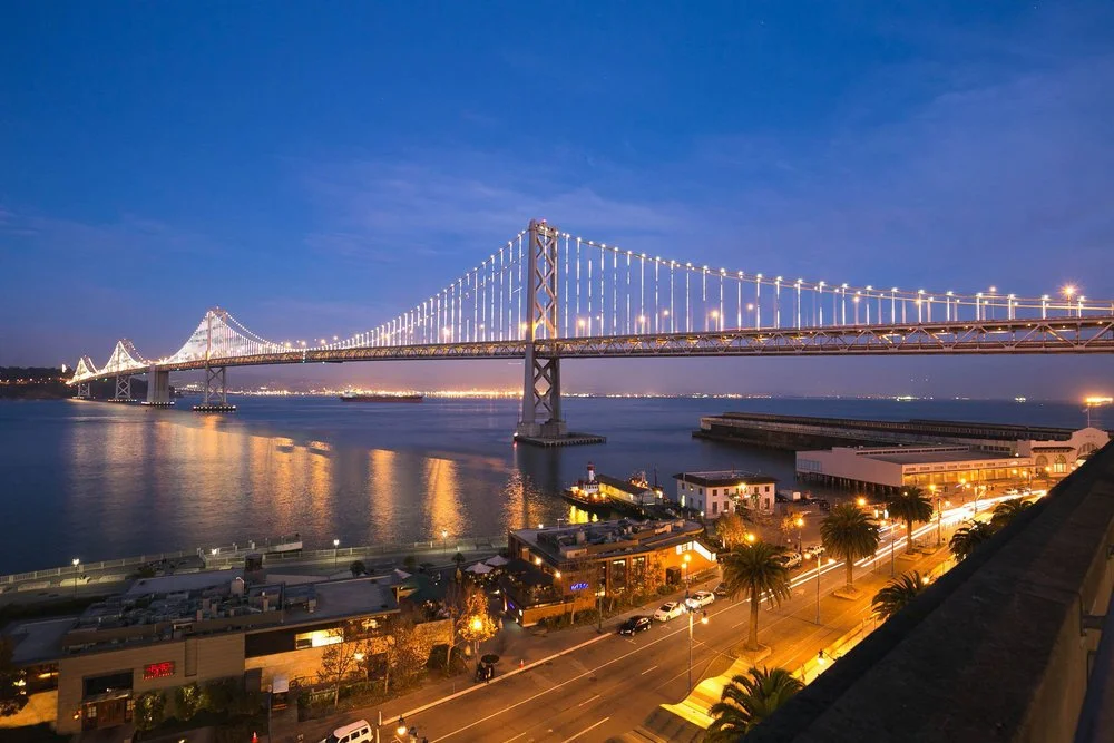 A lit-up suspension bridge at night over a body of water, with city lights reflecting on the water and a road with cars and palm trees in the foreground.
