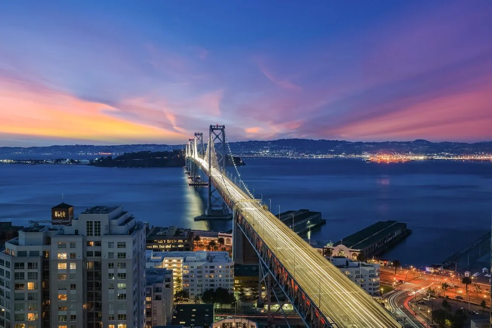 Aerial view of a suspension bridge at sunset over a bay, with city buildings in foreground and rugged hills in the background.