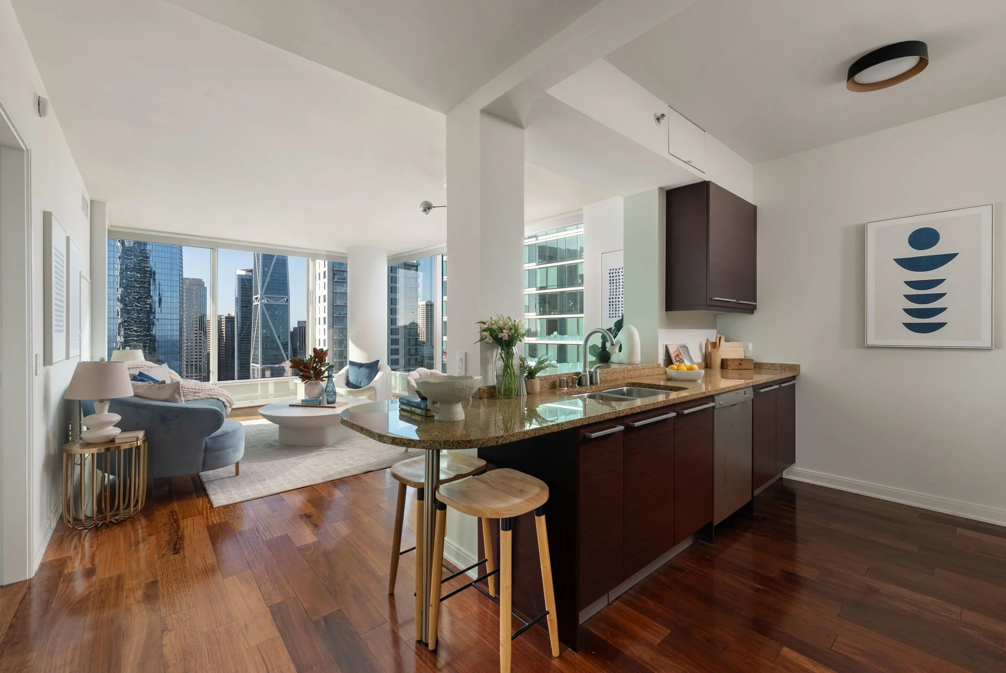 Modern open-plan living room and kitchen with large windows showcasing a city skyline. The kitchen has dark wood cabinetry, a granite countertop, and bar stools. The living area has a blue armchair, a white coffee table, and decorative plants.